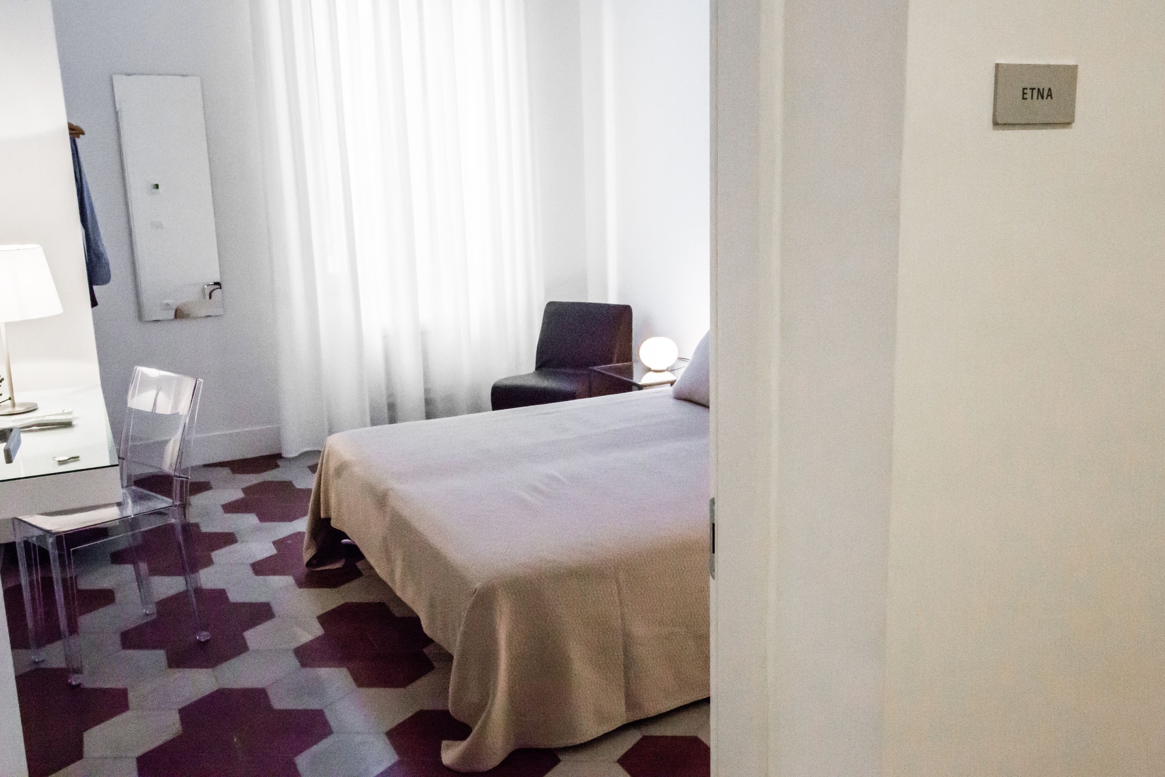 A hotel room with a bed, a desk with a chair, a lamp, and a mirror on the wall.
