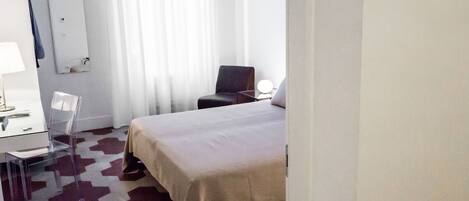 A hotel room with a bed, a desk with a chair, a lamp, and a mirror on the wall.