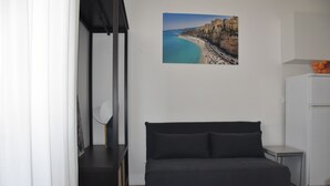 A modern room with a dark-colored sofa, a small side table, a white refrigerator, and a framed coastal landscape picture on the wall. - Rhegion B&B (Reggio Calabria)