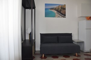 Una habitación moderna con un sofá de color oscuro, una pequeña mesita auxiliar, un refrigerador blanco y un cuadro enmarcado con un paisaje costero en la pared.