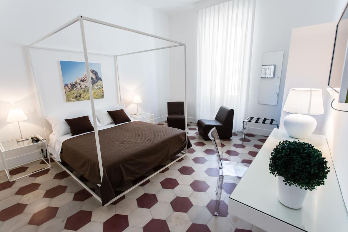 A modern hotel room with a four-poster bed, a desk with a lamp, a chair, and a potted plant.