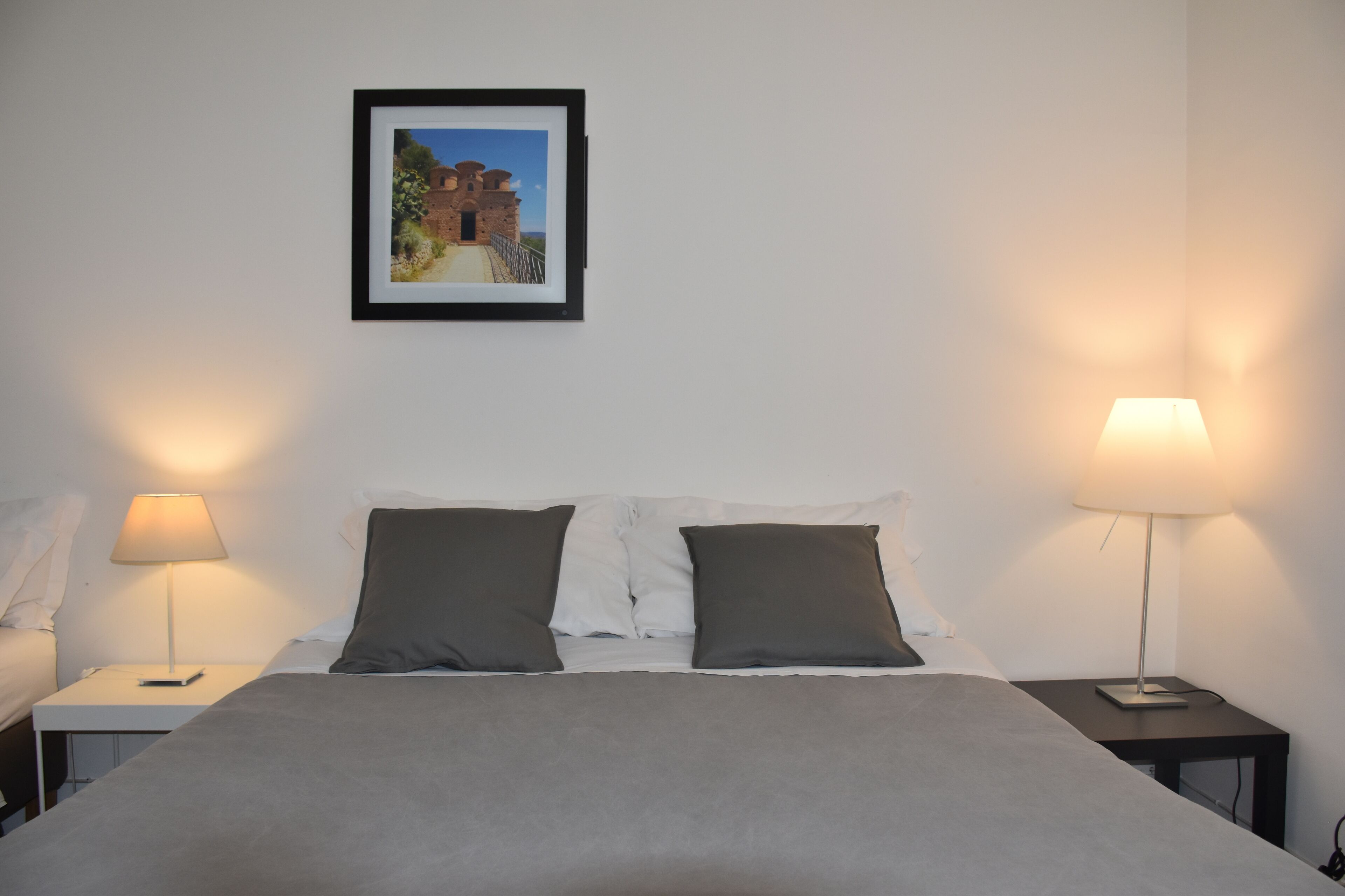 Un letto rifatto con cura, con una coperta grigia e due cuscini, affiancato da due comodini con lampade, uno per lato.