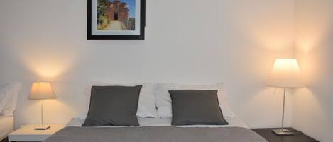 Uma cama bem arrumada com um cobertor cinza e duas almofadas, com duas mesas de cabeceira e abajures, um de cada lado.