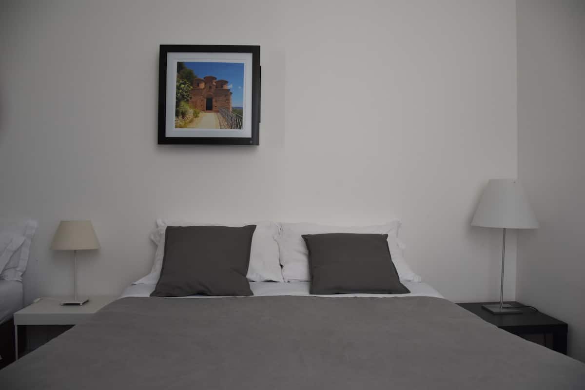 A neatly made bed with a grey blanket and two dark pillows, a framed picture on the wall, and two bedside lamps on either side of the bed.