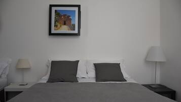 A neatly made bed with a grey blanket and two dark pillows, a framed picture on the wall, and two bedside lamps on either side of the bed.