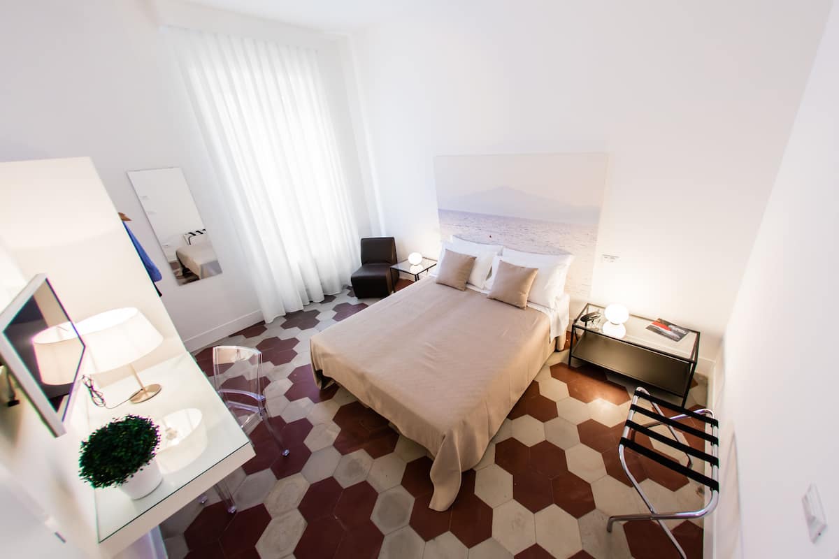 A modern hotel room with a large bed, a bedside table with a lamp, a mirror, a small desk, and a chair.