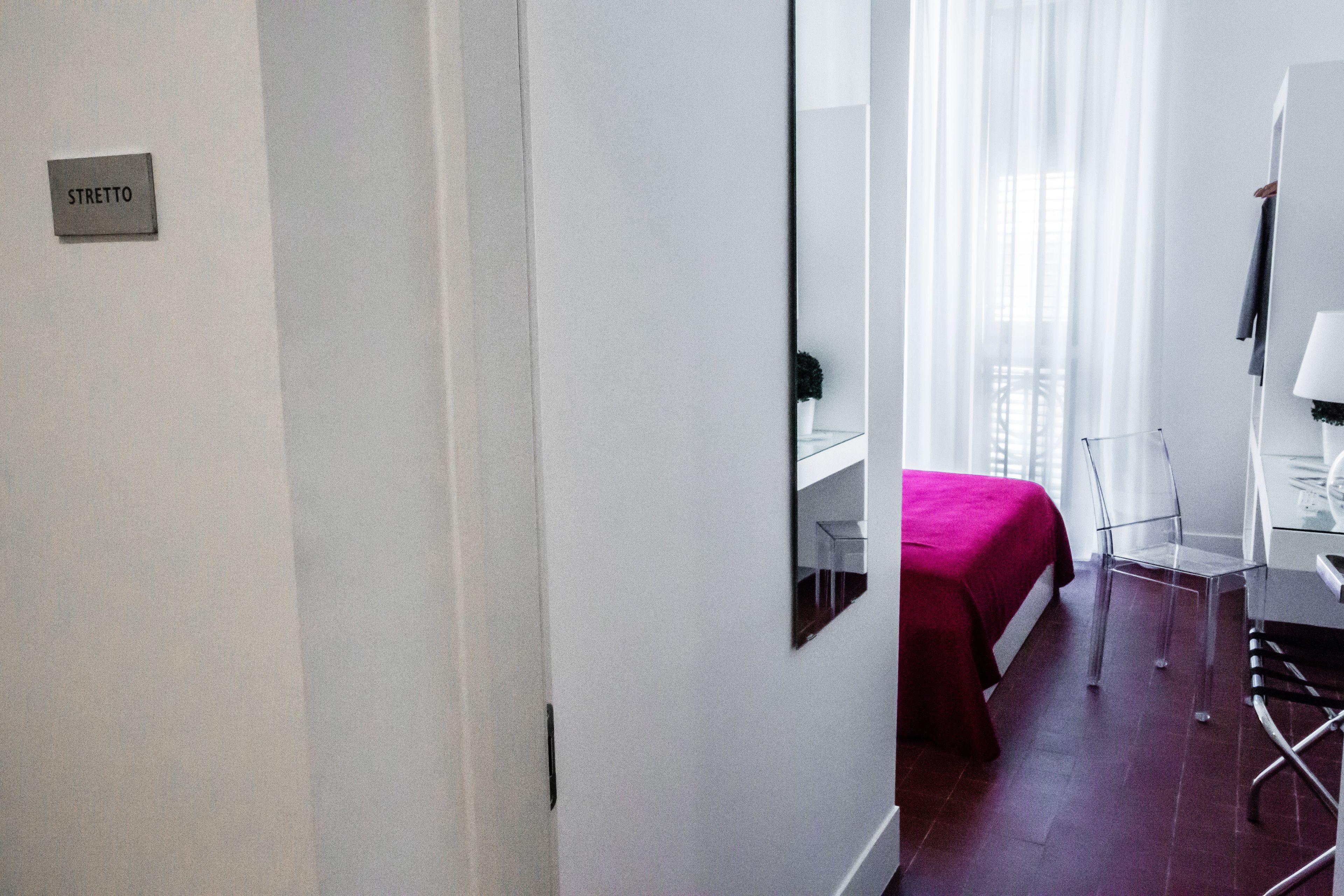 Habitación de hotel con cama, escritorio, silla, espejo y ventana con cortinas translúcidas.