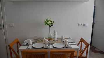 A dining area with a white table set for four, surrounded by wooden chairs. A vase with flowers is placed on the table, and there are various dining utensils and plates.