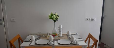 A dining area with a white table set for four, surrounded by wooden chairs. A vase with flowers is placed on the table, and there are various dining utensils and plates.