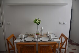 Un comedor con una mesa blanca dispuesta para cuatro personas, rodeada de sillas de madera. Un jarrón con flores está sobre la mesa, y hay cubiertos y platos varios.