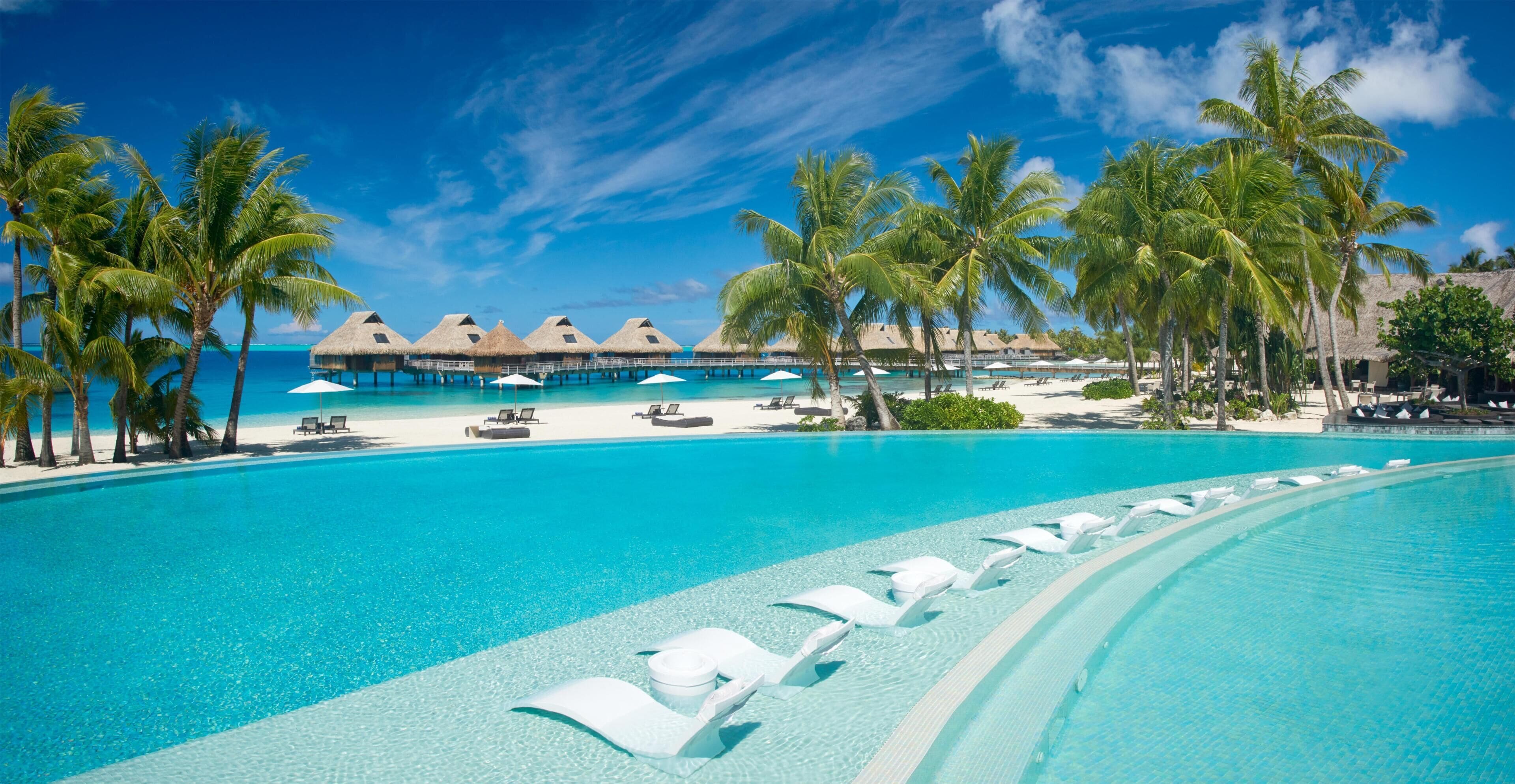 A resort pool with clear blue water, white lounge chairs, palm trees, and overwater bungalows.