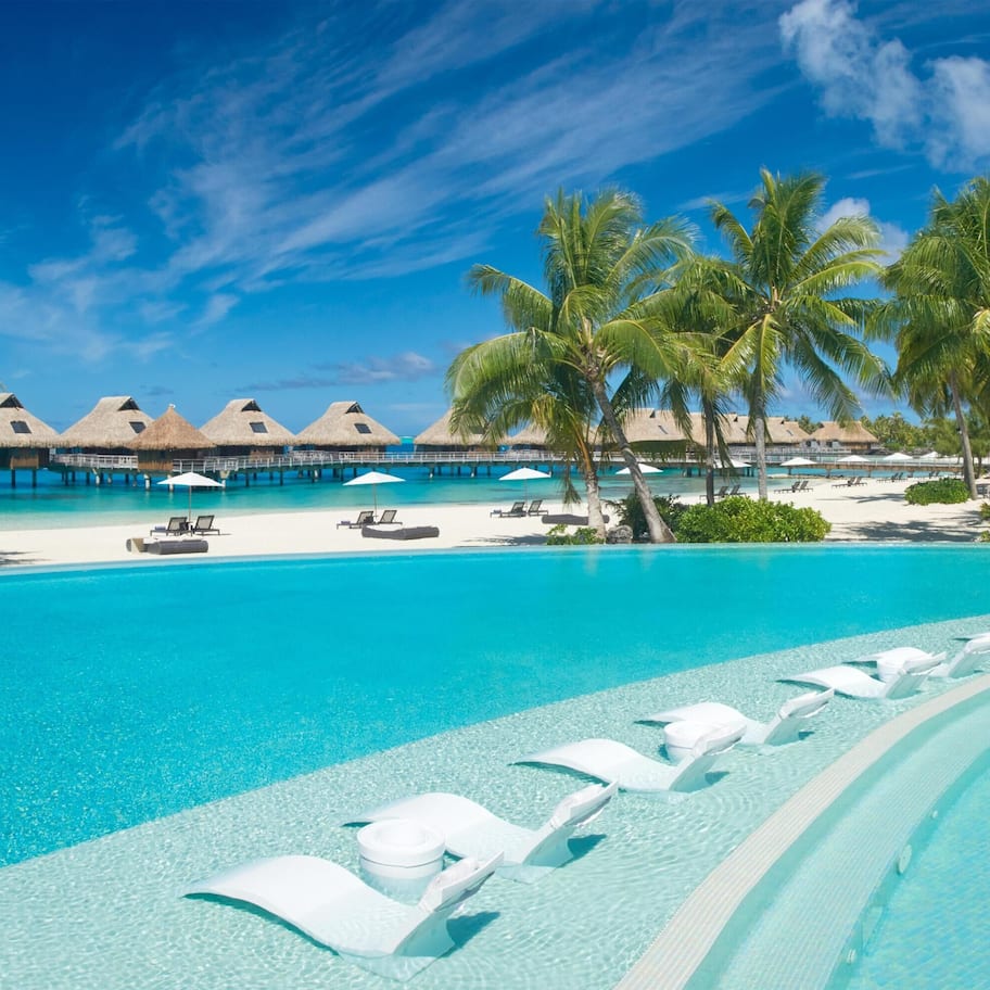 A resort pool with clear blue water, white lounge chairs, palm trees, and overwater bungalows.