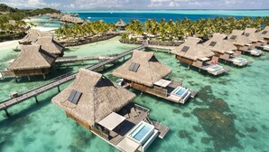 Des bungalows sur pilotis, avec toits de chaume, une eau turquoise cristalline et une passerelle en bois qui les relie.