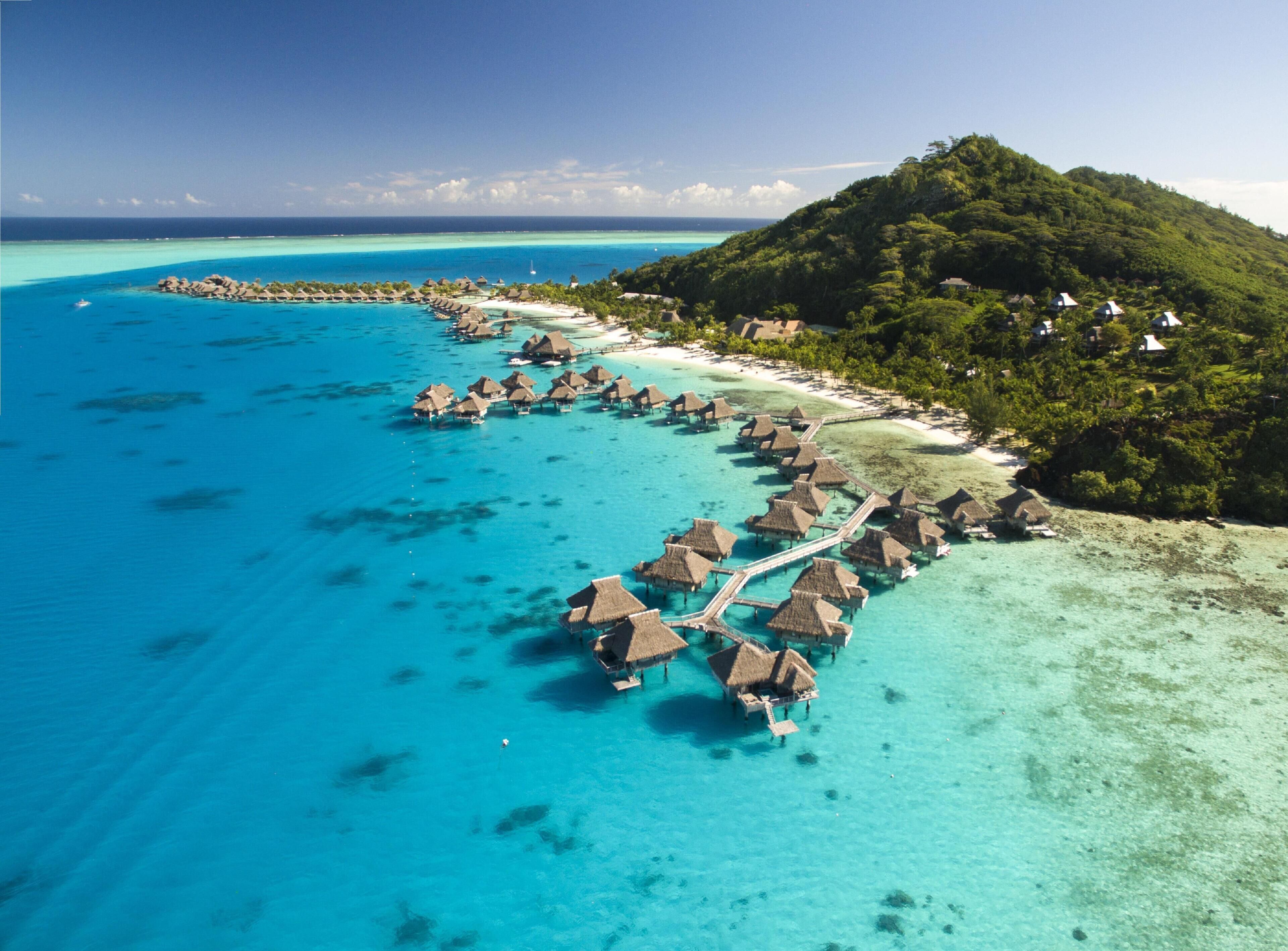 An aerial view of overwater bungalows connected by a walkway, surrounded by turquoise waters and a lush green island.
