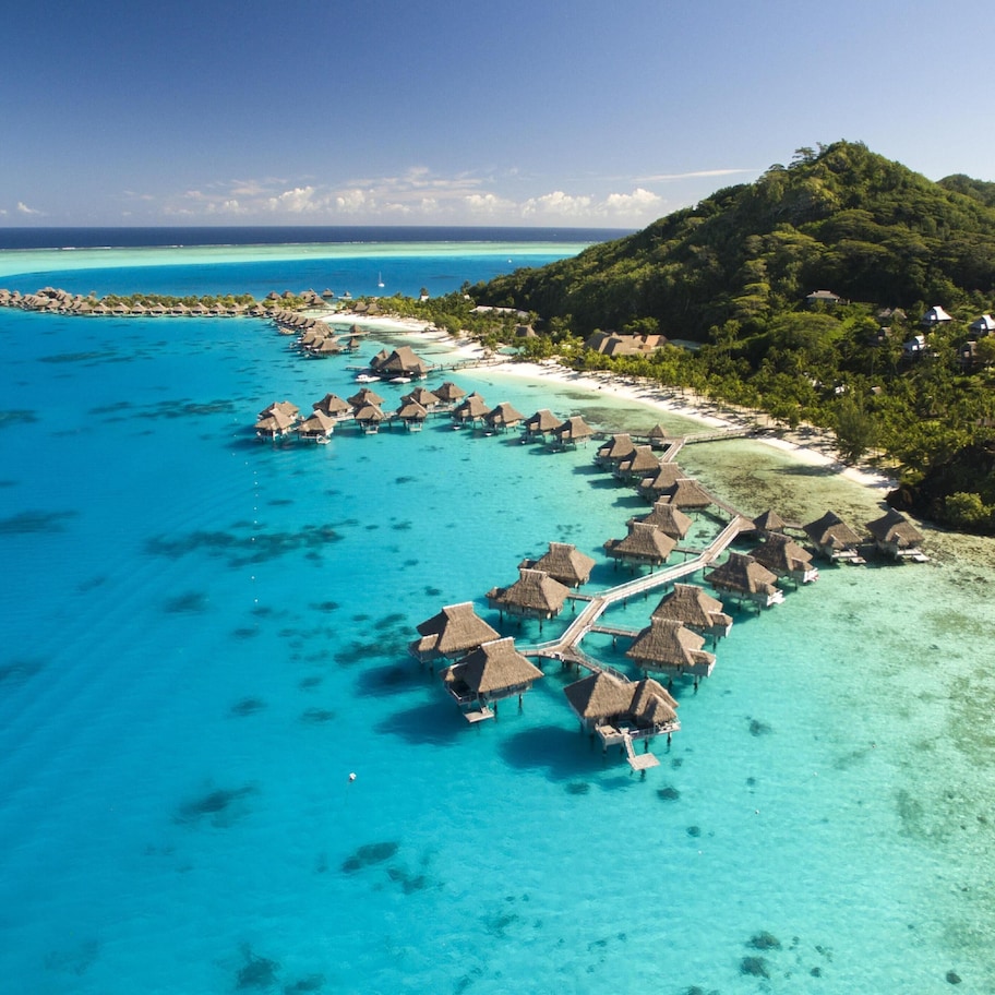 An aerial view of overwater bungalows connected by a walkway, surrounded by turquoise waters and a lush green island.