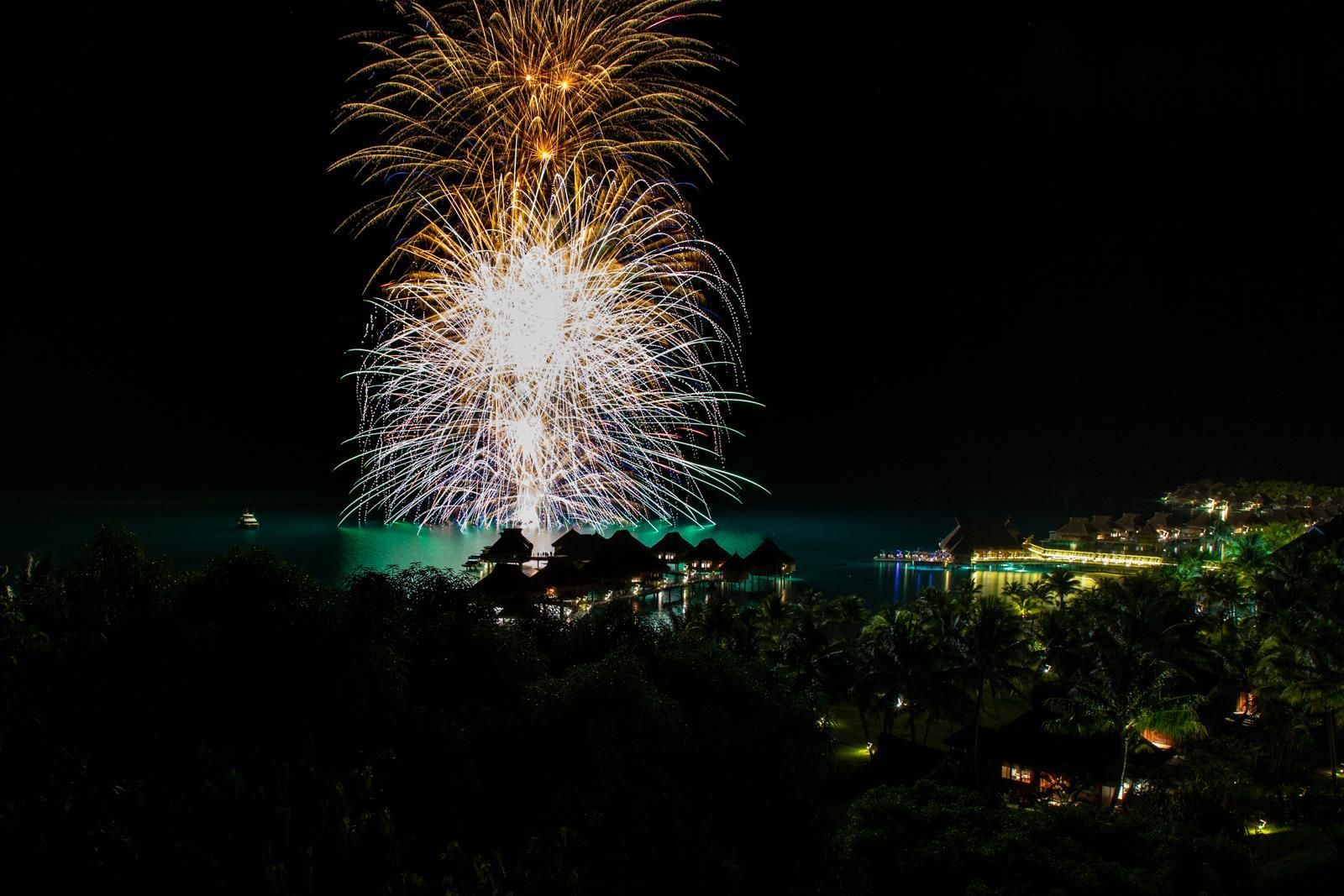 A nighttime view of a resort with overwater bungalows, a boat, and a fireworks display.
