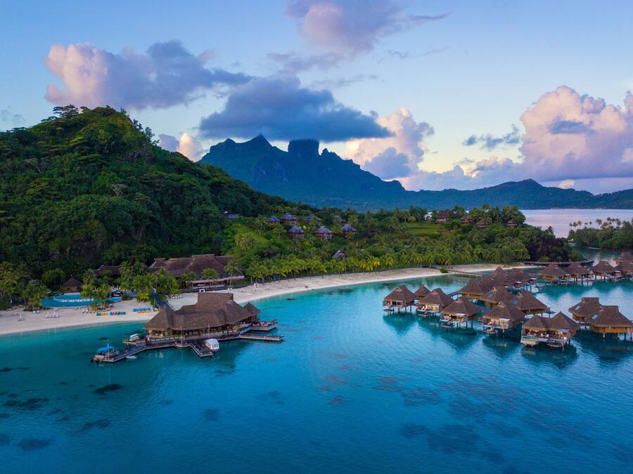 An aerial view of a tropical resort with overwater bungalows, a beach, and lush greenery.