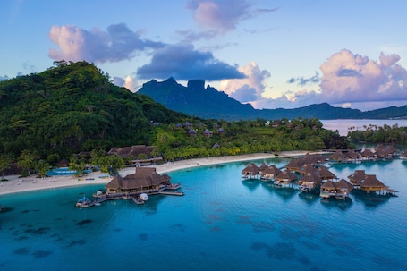 An aerial view of a tropical resort with overwater bungalows, a beach, and lush greenery.