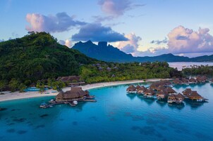 Vista aérea de un resort tropical con bungalows sobre el agua, una playa y abundante vegetación.