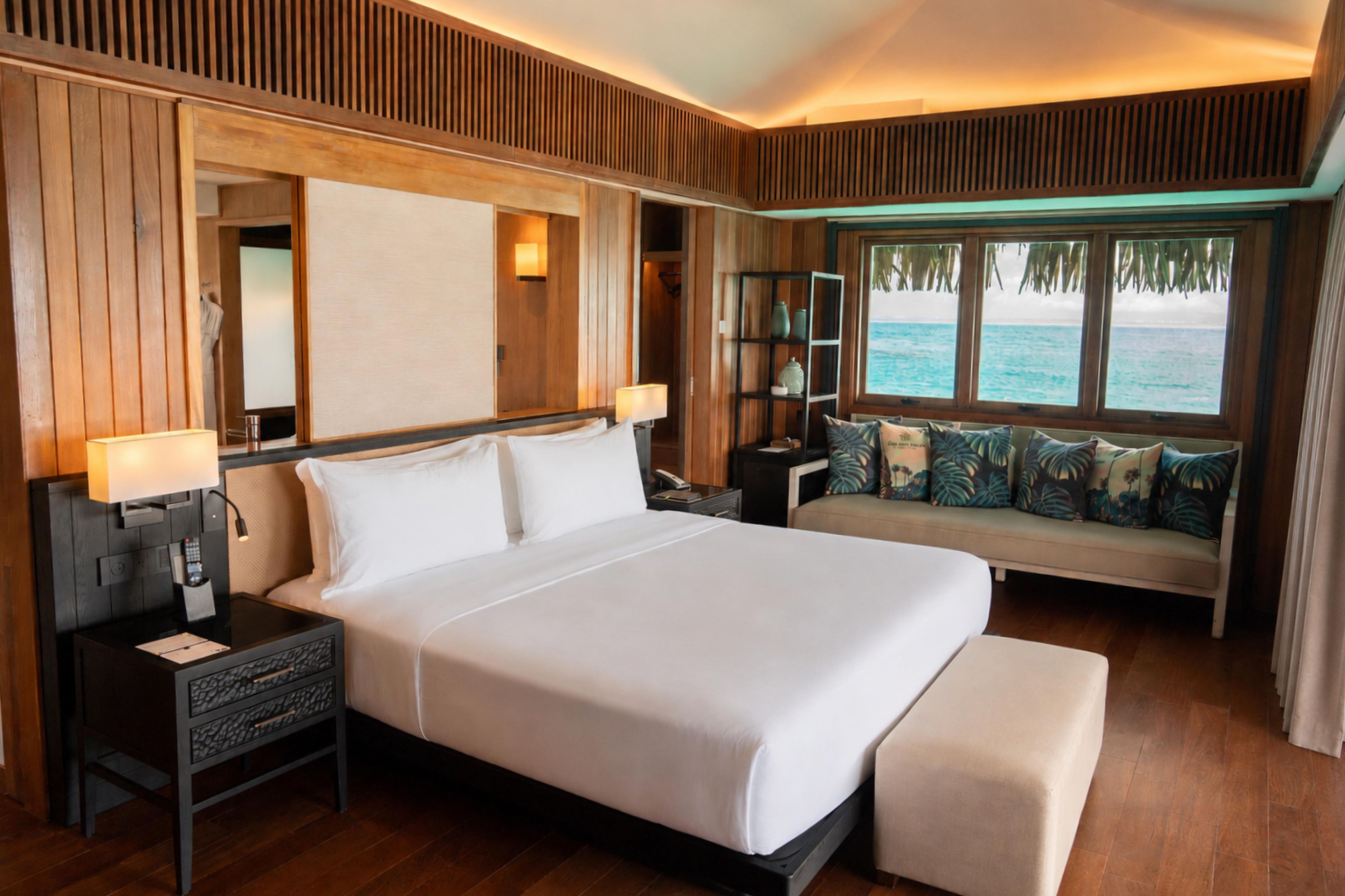 A hotel room with a large bed, bedside tables, a nightstand, a sofa with cushions, a window with a view of the sea, and wooden paneling on the walls.
