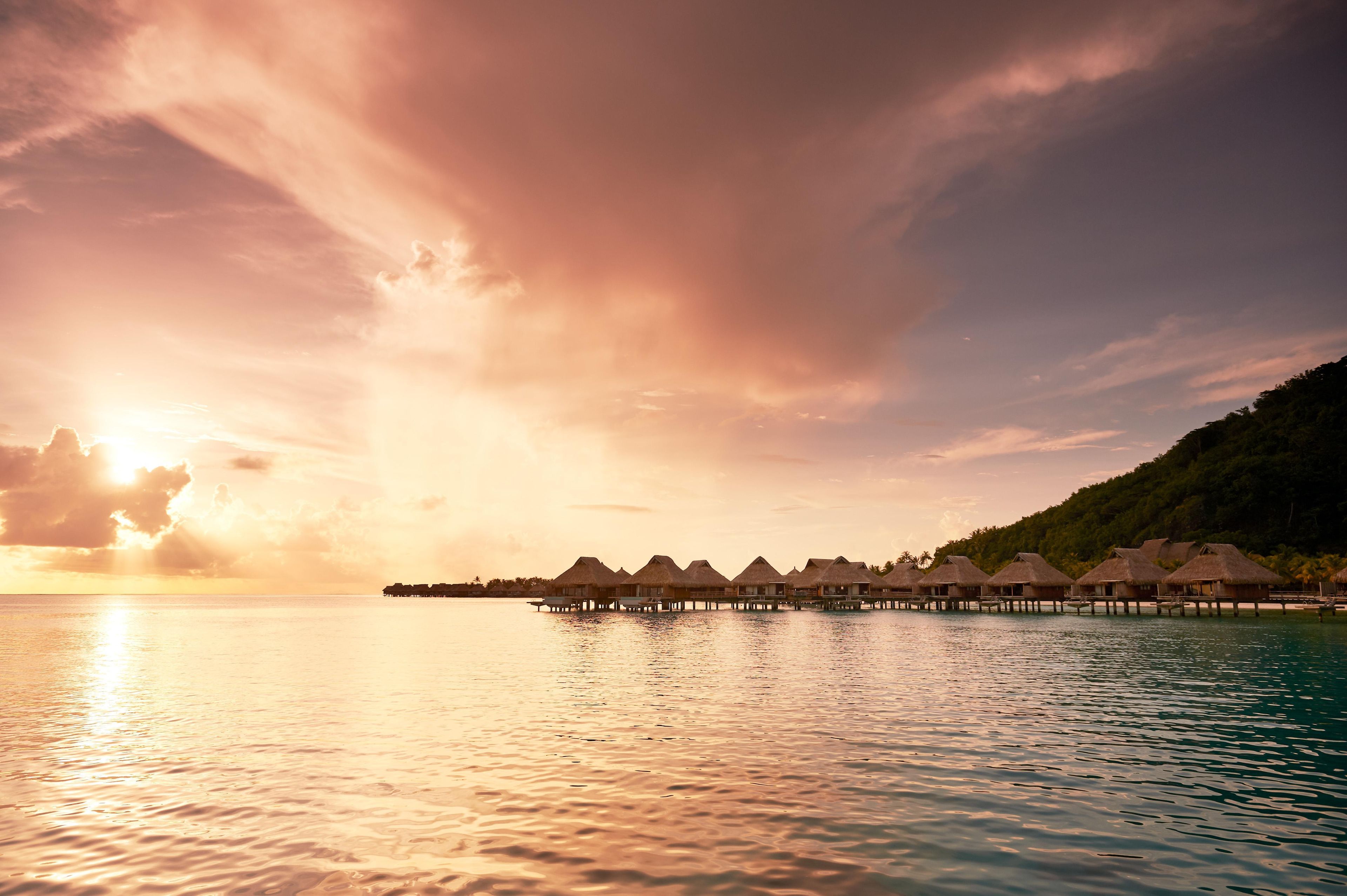 Overwater bungalows with thatched roofs, connected by walkways, and a calm sea at sunset.