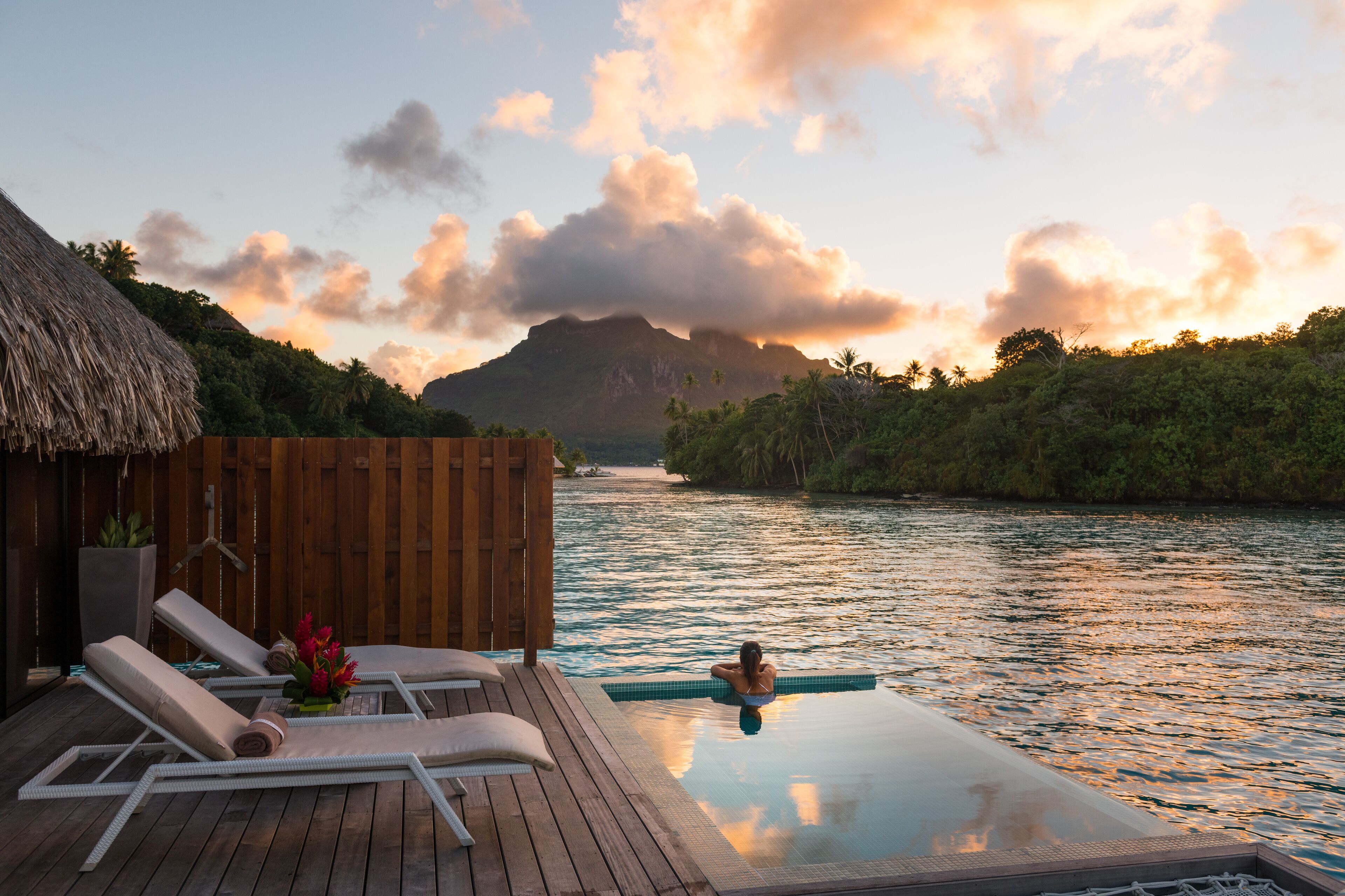 Un terrazzo in legno con poltrone relax e una piscina, con vista su un corpo d'acqua e una montagna sullo sfondo.
