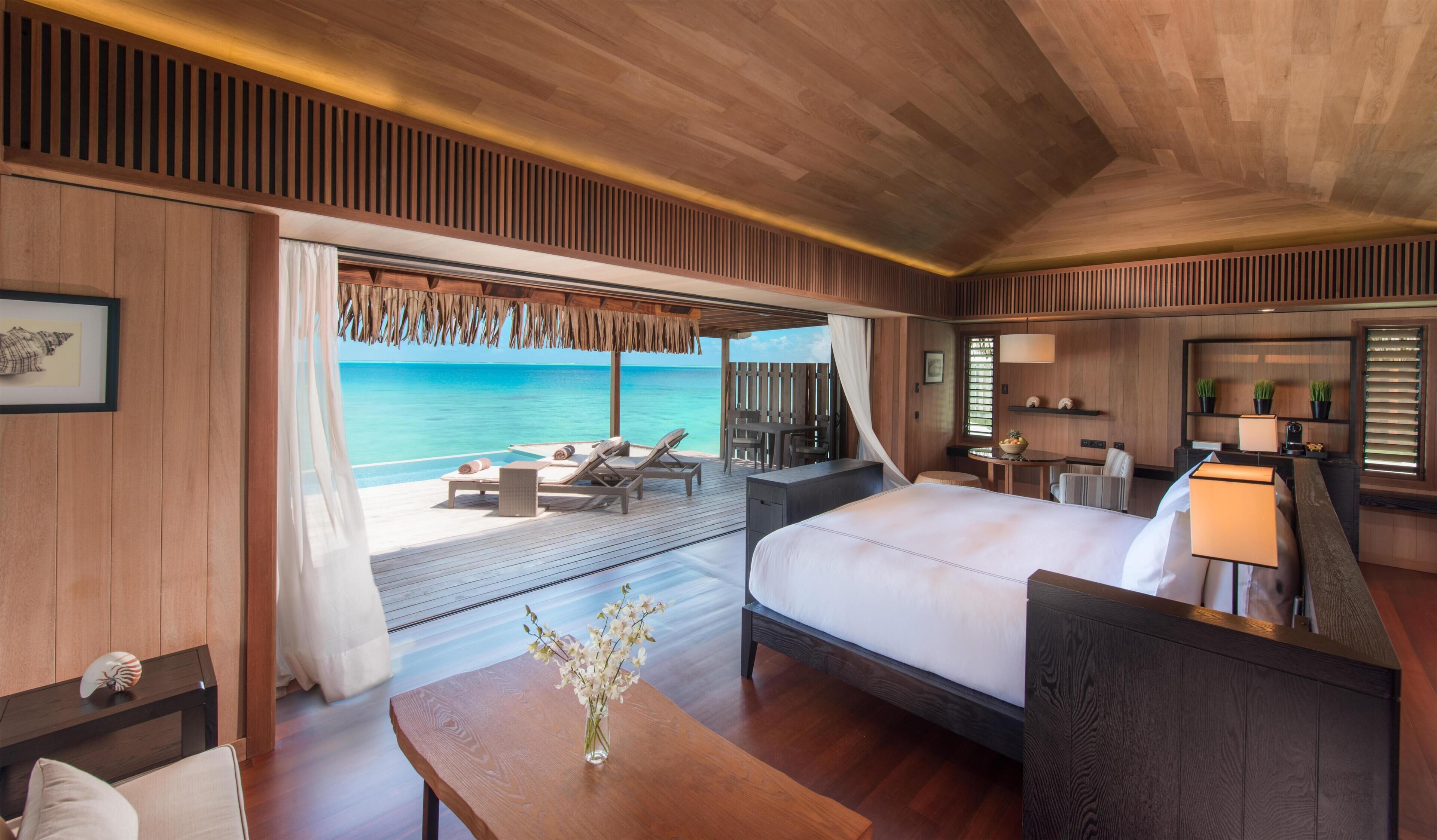 A hotel room with a bed, a desk, a chair, a small table with a vase of flowers, and a view of the ocean.