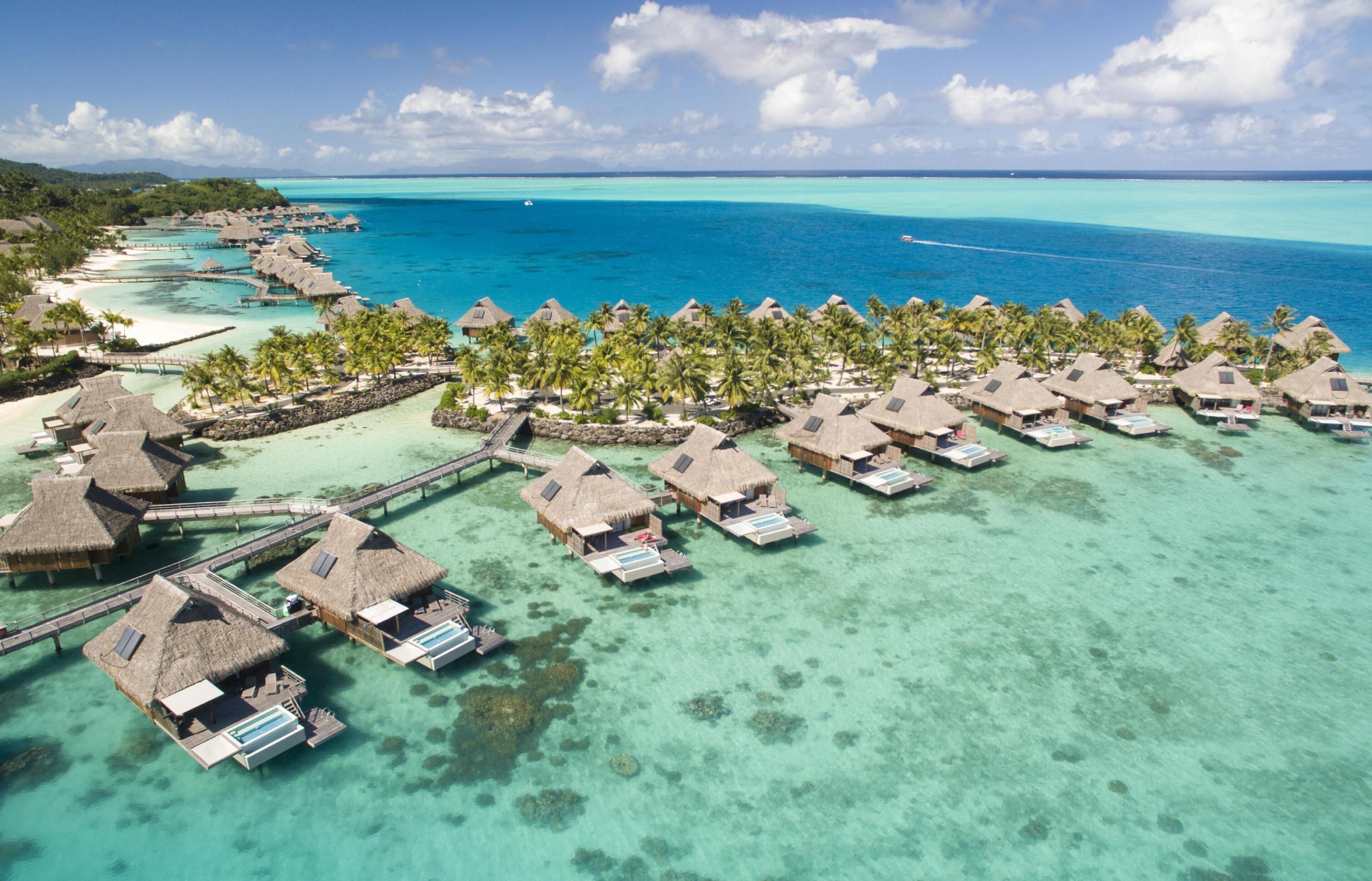 An aerial view of overwater bungalows with thatched roofs, connected by walkways, set against a turquoise sea and clear sky.