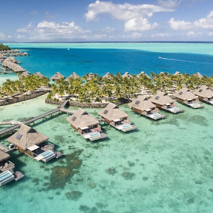An aerial view of overwater bungalows with thatched roofs, connected by walkways, set against a turquoise sea and clear sky.
