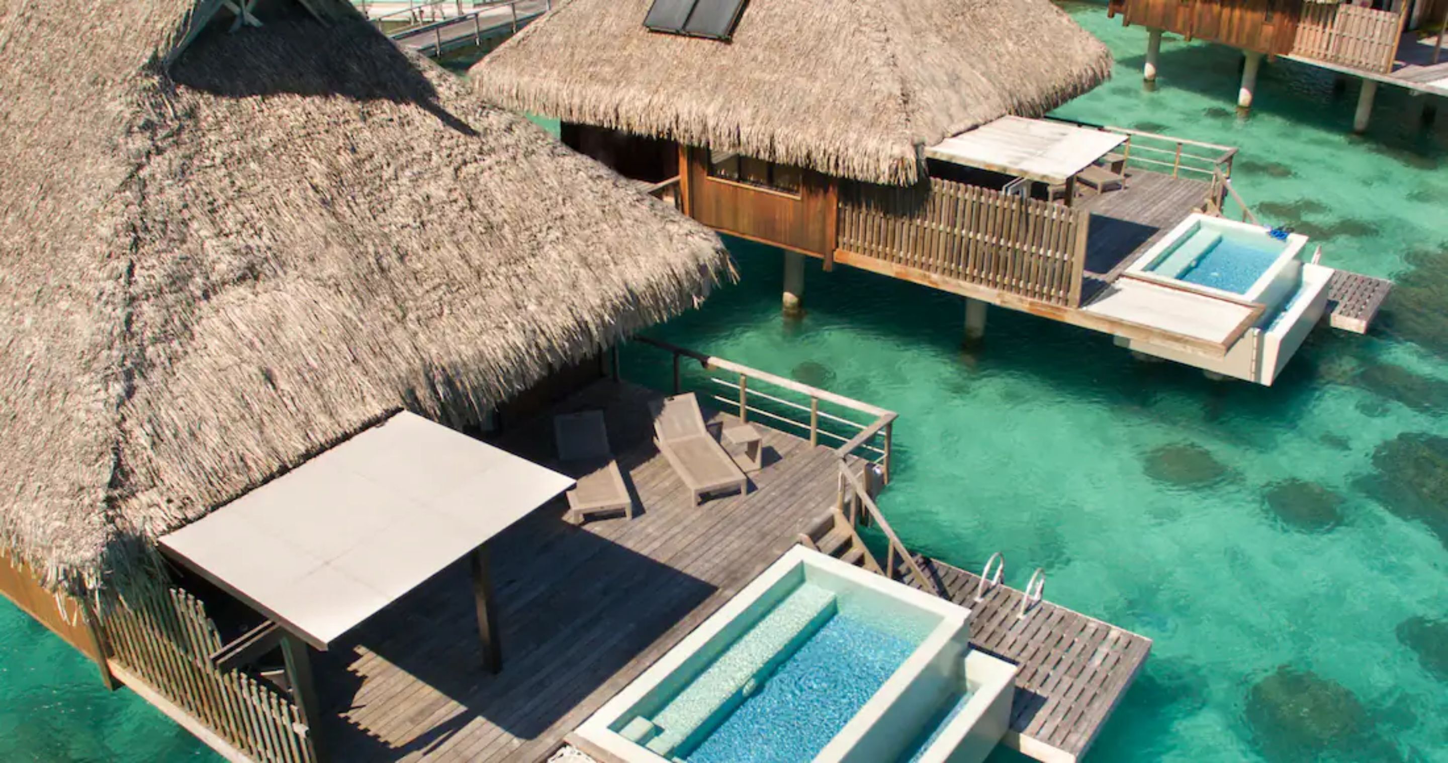 An overwater bungalow with a thatched roof, a pool, and wooden deck furniture.