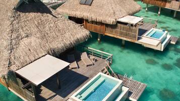 An overwater bungalow with a thatched roof, a pool, and wooden deck furniture.
