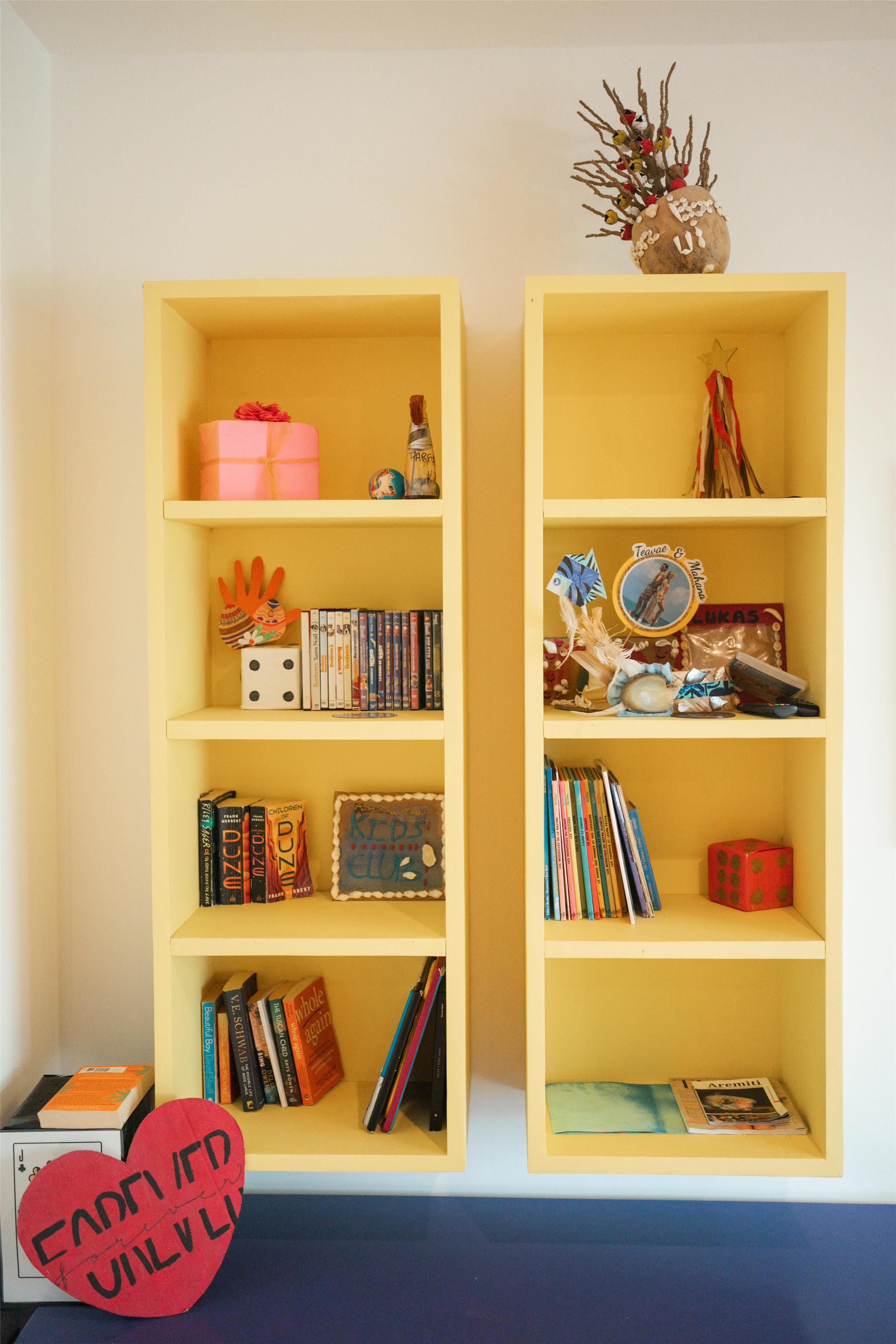 Two yellow bookshelves with various books, a red heart-shaped object, and a small red gift box.