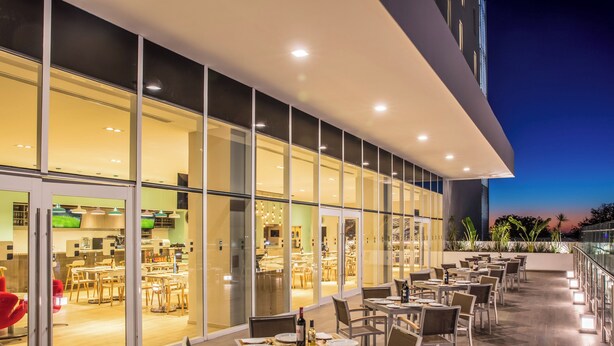 Un área de comedor al aire libre con mesas y sillas, un muro de vidrio que separa el comedor de la cocina, y vistas del horizonte urbano al atardecer.