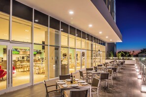 Un espace de restauration en plein air avec des tables et des chaises, une paroi en verre séparant l’espace de restauration de la cuisine, et une vue sur les toits de la ville au crépuscule.