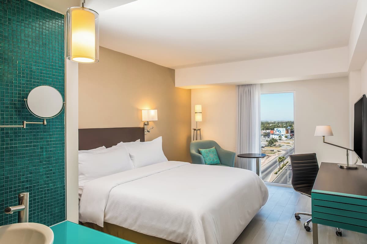 A hotel room with a large bed, a desk, a chair, a TV, a window with a view, and a bathroom with a mirror and sink.