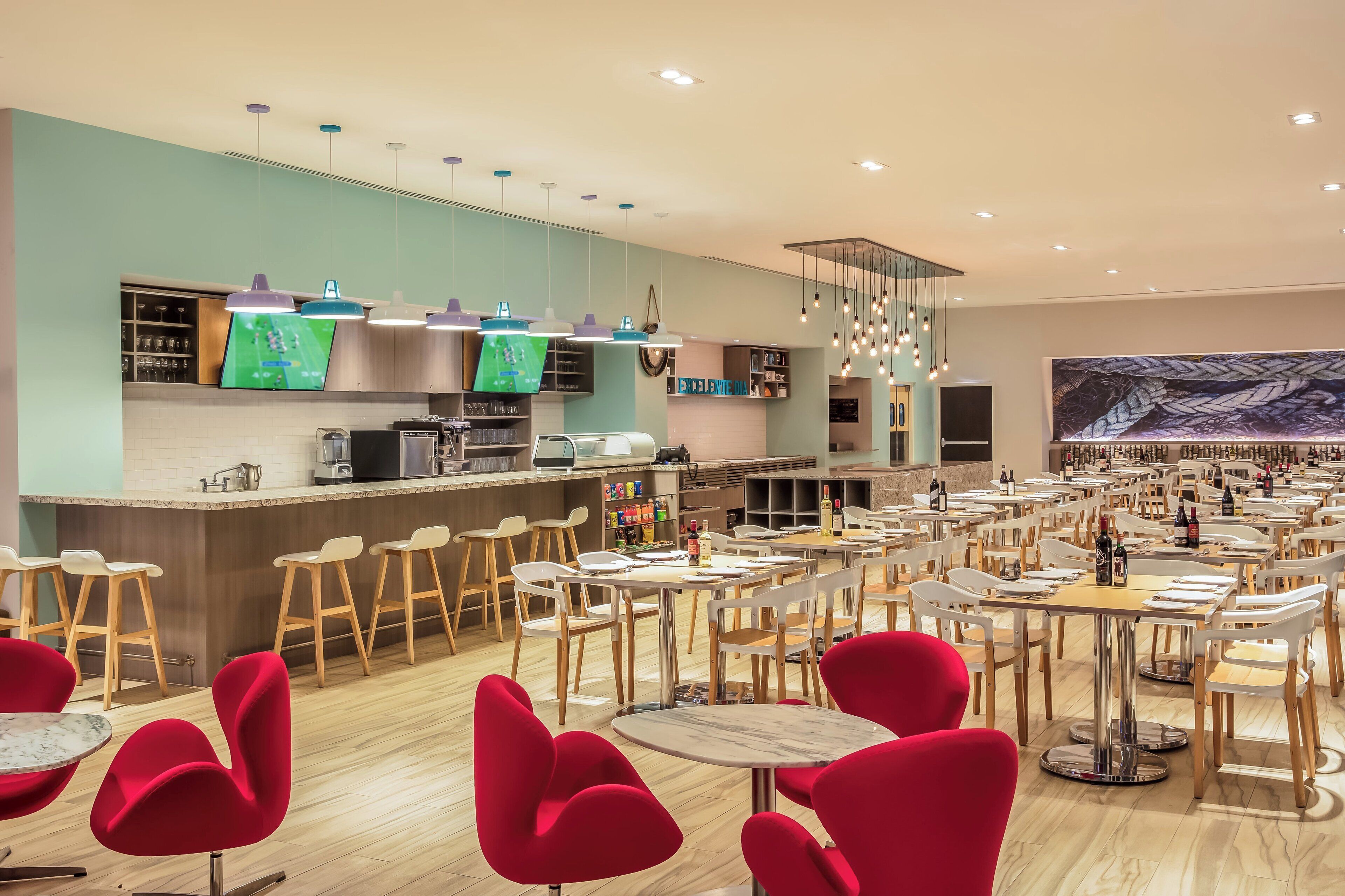 A modern restaurant with a bar area, multiple tables and chairs, and a large screen displaying a game.