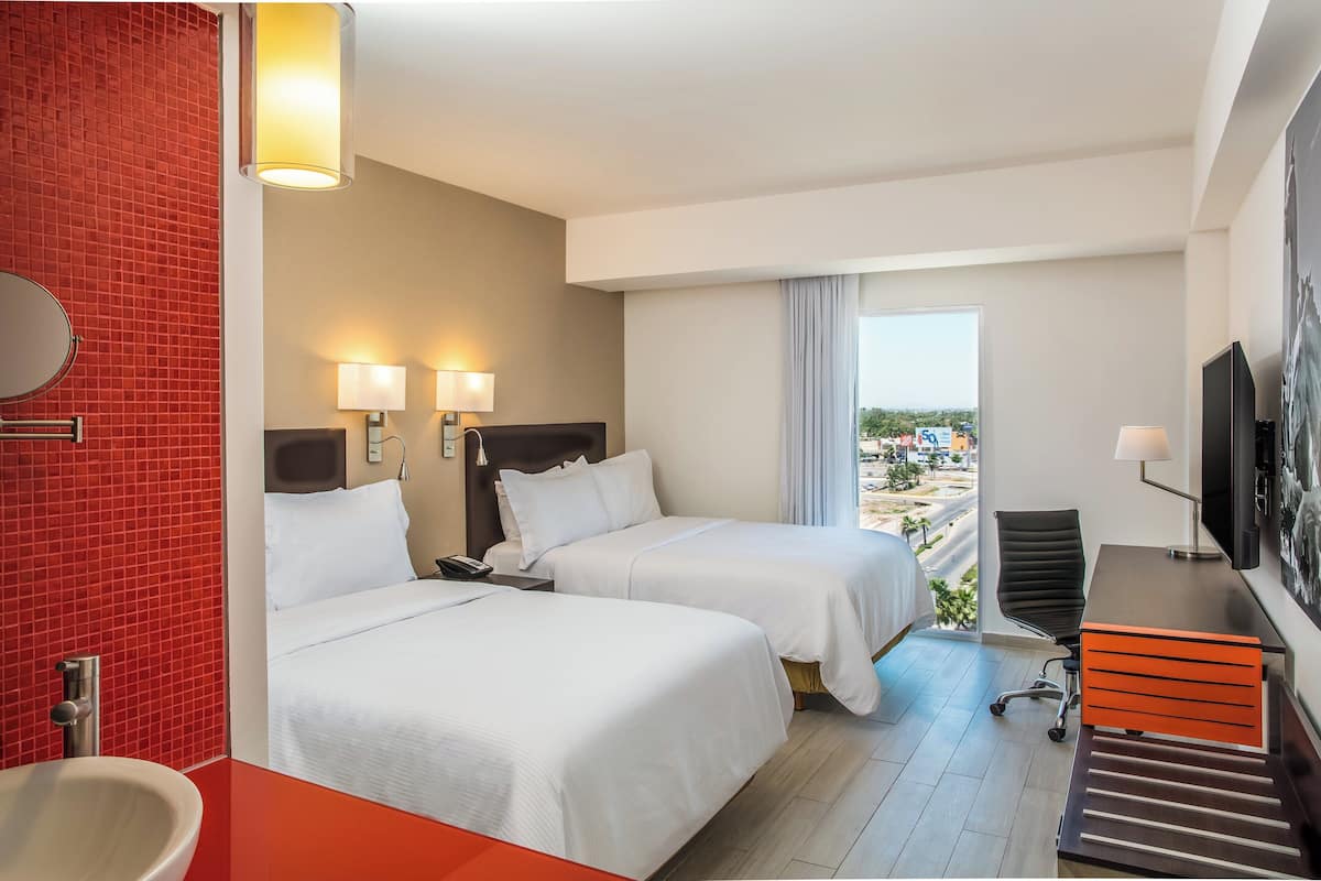 A hotel room with two beds, a desk, a chair, a TV, and a bathroom with a red-tiled wall and a sink.