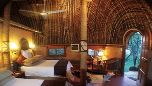 A room with two beds, a wooden table, a chair, a fireplace, and a large arched doorway leading outside. - aha Shakaland (Eshowe)