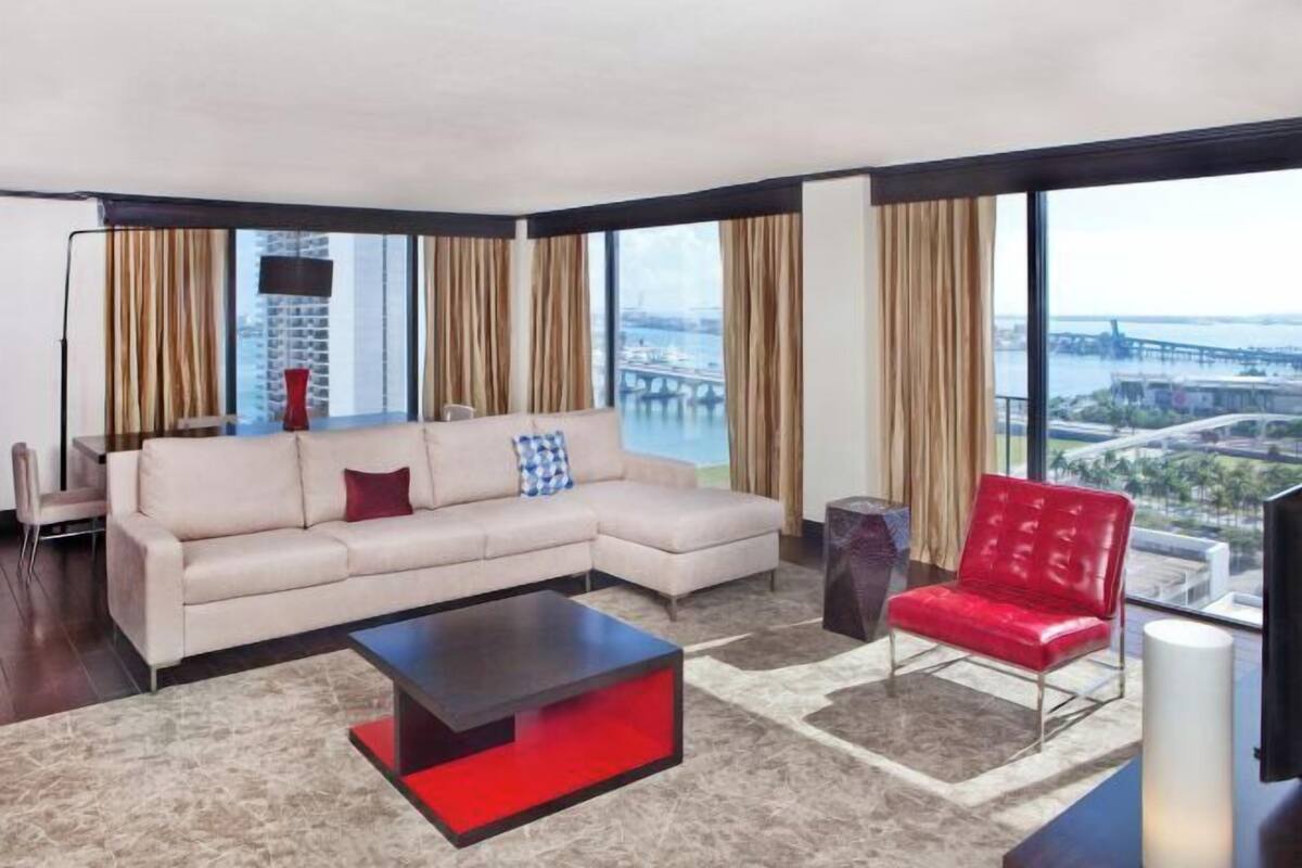 A modern living room with a beige sectional sofa, a red chair, a black coffee table, and large windows offering a view of a bridge and cityscape.
