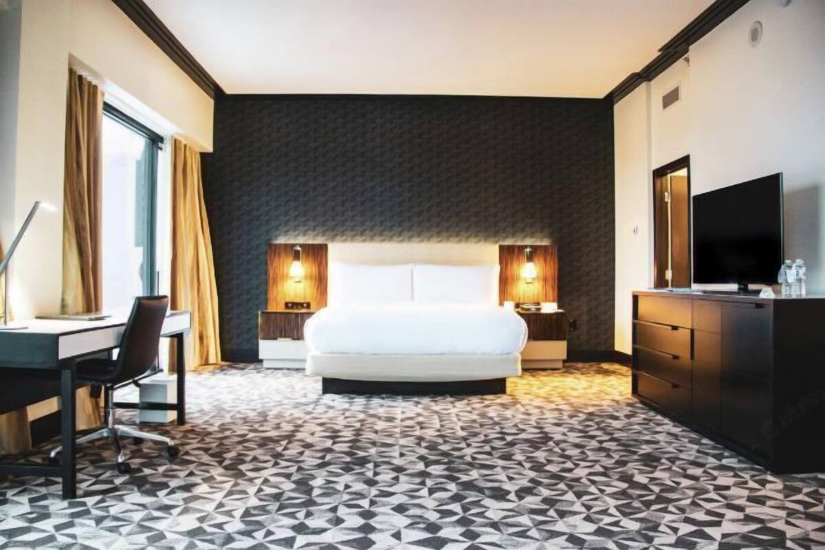 A hotel room with a bed, desk, chair, TV, and a patterned floor.