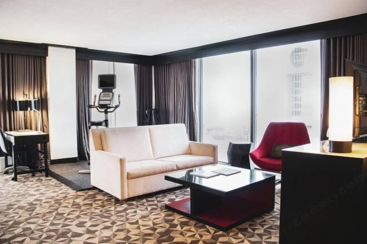 A modern hotel room with a beige sofa, a red armchair, a glass coffee table, and a large window offering a city view.