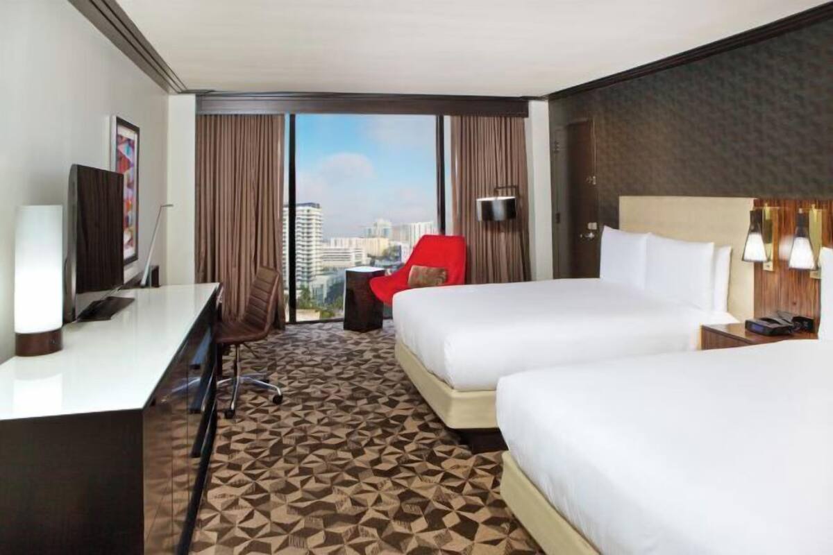 A hotel room with two beds, a desk, a chair, a view of the city, and a large window.