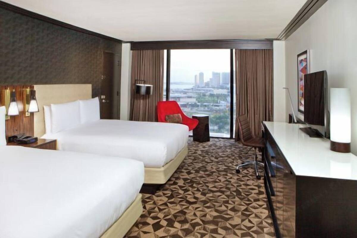 A hotel room with two beds, a desk, a chair, a TV, and a large window with a city view.