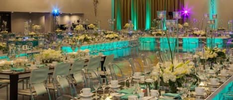 Un salón de banquetes con mesas largas dispuestas para un evento formal, con sillas transparentes y centros de mesa florales.