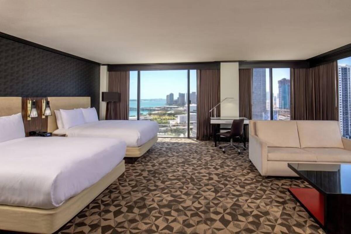 A hotel room with two beds, a desk, a sofa, and large windows with a city view.
