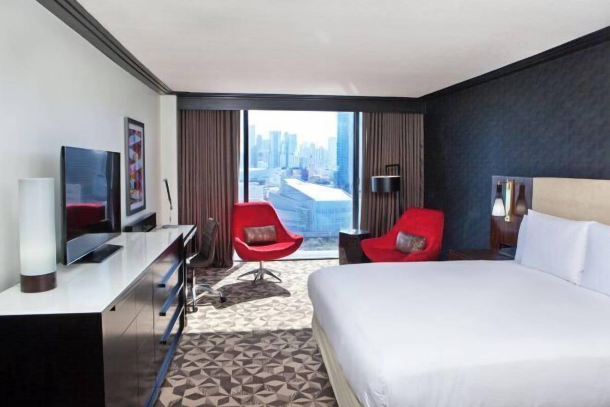 A hotel room with a large bed, a desk with a TV, two red chairs, a city view, and a patterned carpet.