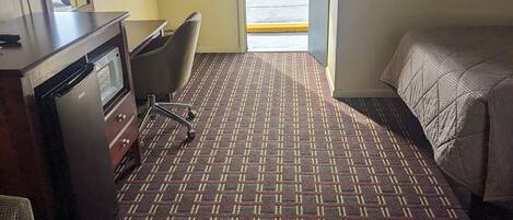 A hotel room with a patterned carpet, a bed, a desk with a lamp, a chair, and a view of the outside.