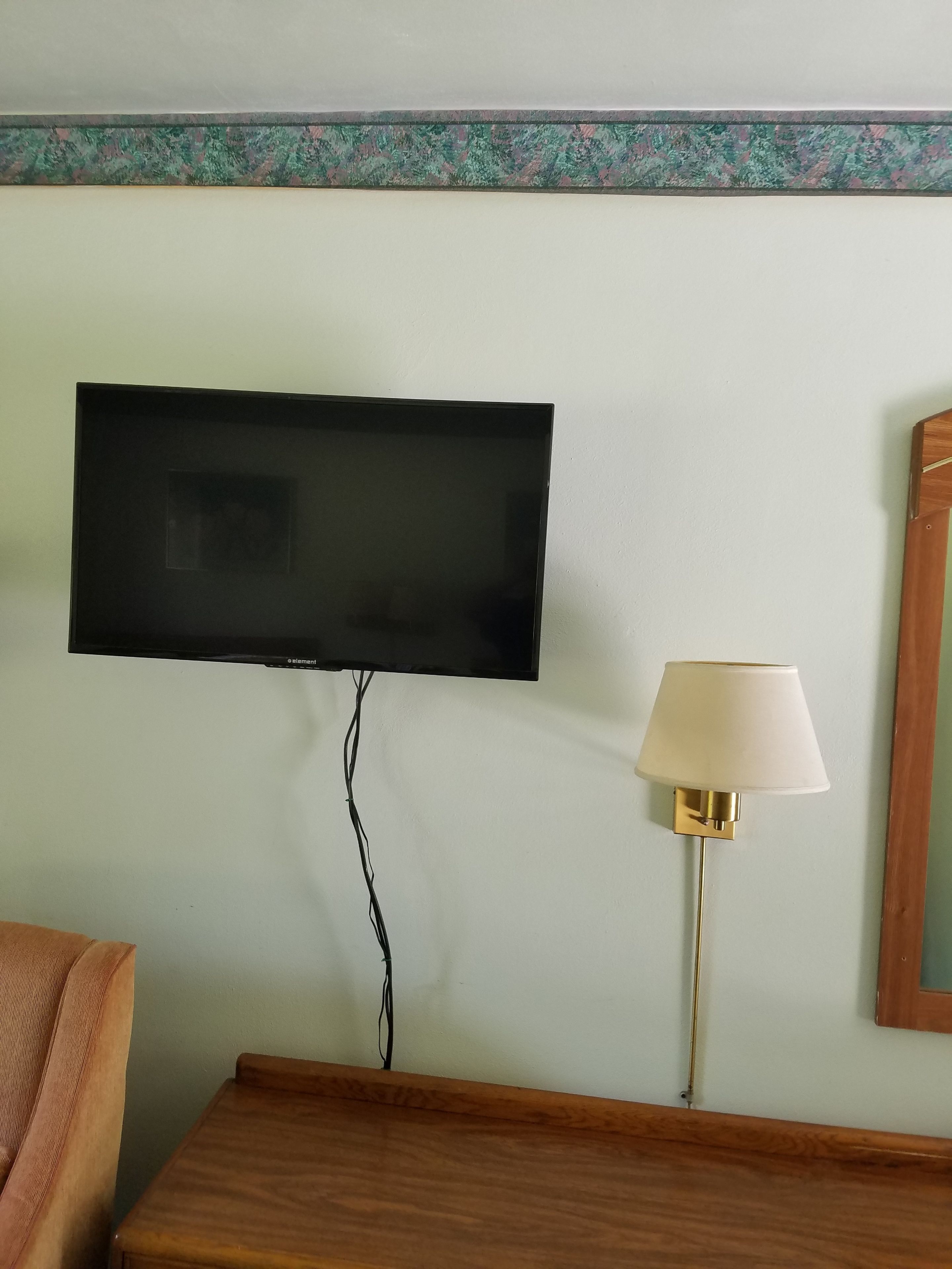 En platt-TV som är monterad på väggen, en sänglampa och ett litet bord i trä som står vid sängen.