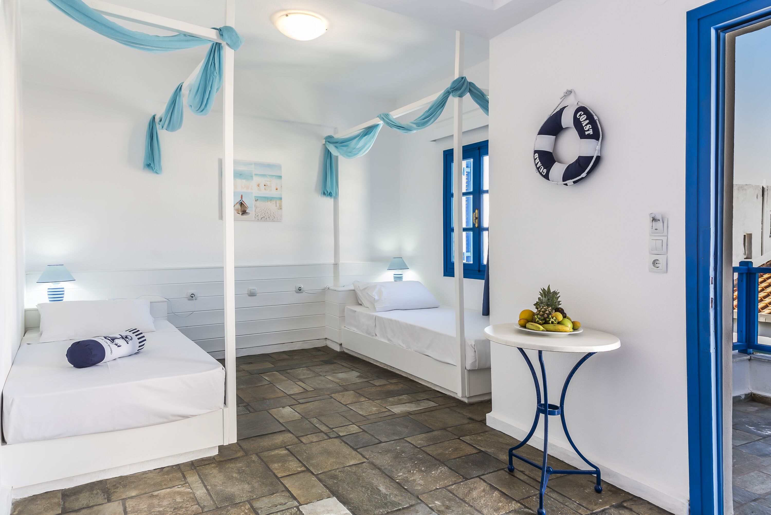 Una habitación con una cama, una mesita con fruta, un flotador en la pared y una puerta azul.