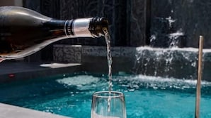 A hand pouring champagne into a glass by a pool with a waterfall feature in the background. - Elmi Suites Beach Hotel - All Inclusive (Hersonissos)